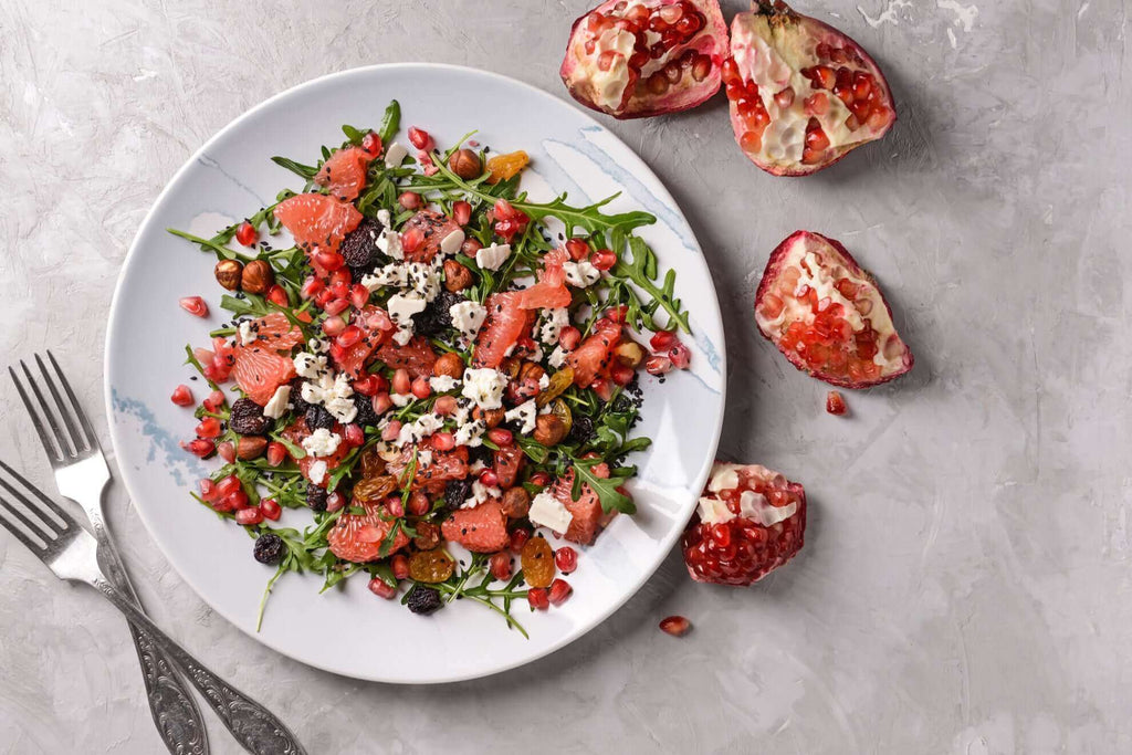 Colorful arugula salad topped with pomegranate seeds, citrus, and feta cheese, served on a white plate.