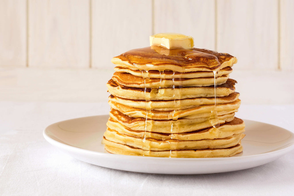 Stack of golden pancakes drizzled with syrup, topped with a pat of butter on a cream plate.