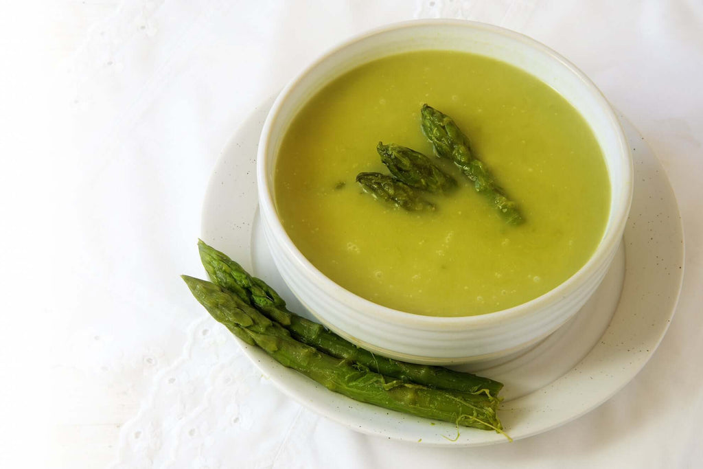 Bowl of creamy asparagus soup garnished with fresh asparagus tips, served on a white plate with additional asparagus.