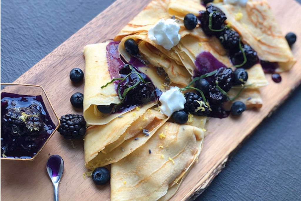 Close-up of delicious crepes topped with berries, lavender jam, and whipped cream on a wooden platter.