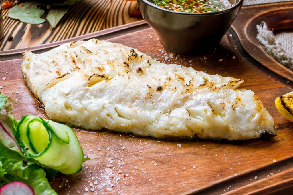 Grilled ling cod fillet served on a wooden board with fresh greens and a dipping sauce.