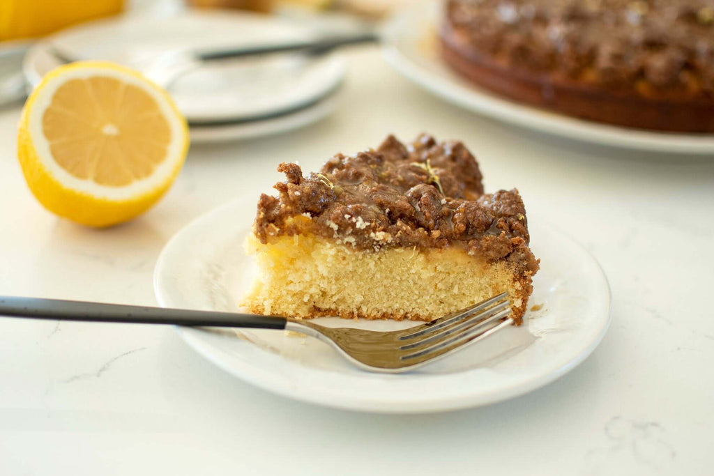 Slice of Meyer lemon coffee cake on a plate with a fork, garnished with a lemon half in the background.