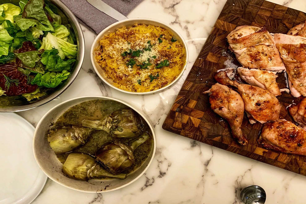 Delicious meal featuring grilled chicken, artichokes, creamy casserole, and fresh salad on a marble table.
