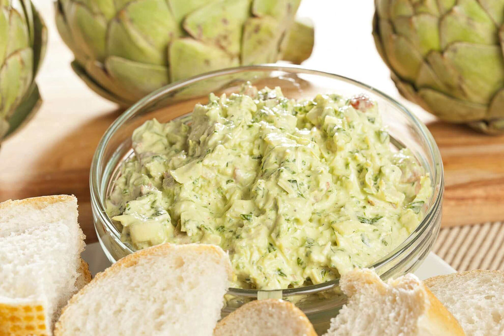 Creamy artichoke dip served in a glass bowl, surrounded by slices of bread and fresh artichokes.