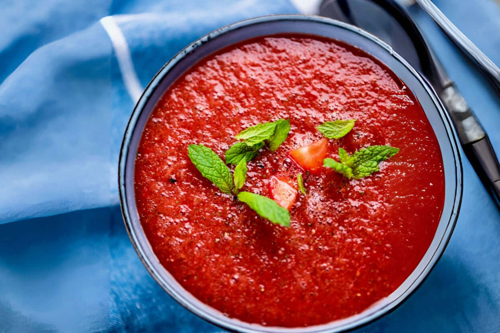 Bowl of fresh tomato watermelon gazpacho garnished with mint and tomato, served on a blue cloth backdrop.