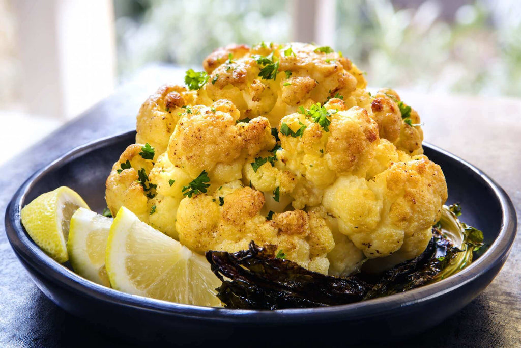 Roasted cauliflower garnished with herbs and served with lemon wedges on a black plate.